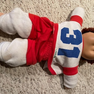 Pode incluir: Uma boneca Cabbage Patch Kid vestindo um jersey branco e vermelho com o número azul "13" na frente. A boneca tem cabelo castanho e uma manga listrada vermelha e branca.