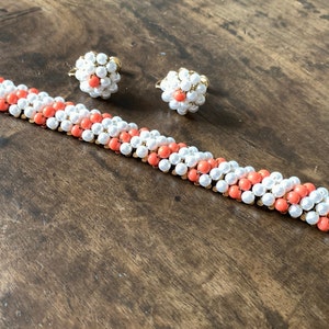Puede incluir: Un conjunto de joyas de coral y perlas blancas que incluye un brazalete y un par de pendientes. El brazalete está hecho de pequeñas cuentas redondas en colores alternos de coral y blanco. Los pendientes también están hechos de pequeñas cuentas redondas en colores alternos de coral y blanco. Las joyas son de color dorado.