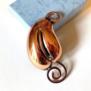 May include: A copper-coloured brooch in the shape of a leaf with a swirl design. The brooch is shiny and has a smooth surface.