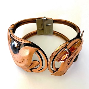 Puede incluir: Un brazalete de cobre con un diseño intrincado que presenta hojas estilizadas y un cierre de plata. El brazalete está hecho de múltiples capas de cobre, creando un aspecto texturizado.