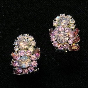 Pode incluir: Um par de brincos vintage de pressão. Cada brinco apresenta um aglomerado de strass roxos claros e rosa em vários tamanhos, com pedras em forma de marquesa roxas mais escuras. Os brincos têm uma moldura de metal prateado.