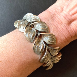 Puede incluir: Un brazalete plateado con un diseño de hojas. El brazalete está hecho de metal y tiene un acabado texturizado.