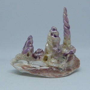 Puede incluir: Una peque&ntilde;a escultura caprichosa hecha de conchas marinas. La escultura presenta un grupo de conchas marinas blancas y moradas dispuestas en una formaci&oacute;n de torre. La escultura est&aacute; colocada sobre una base de una concha marina m&aacute;s grande.