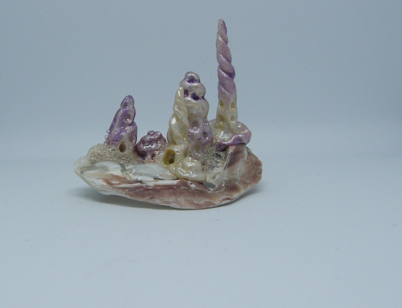 Puede incluir: Una peque&ntilde;a escultura caprichosa hecha de conchas marinas. La escultura presenta un grupo de conchas marinas blancas y moradas dispuestas en una formaci&oacute;n de torre. La escultura est&aacute; colocada sobre una base de una concha marina m&aacute;s grande.