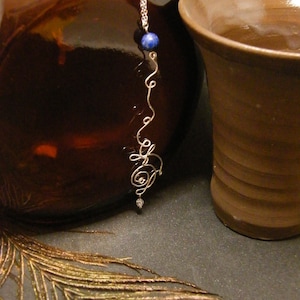 Peut inclure: Un collier en fil d'argent avec un pendentif en pierre bleue et un petit charme en cristal transparent. Le collier est posé sur une surface brune avec une plume de paon.