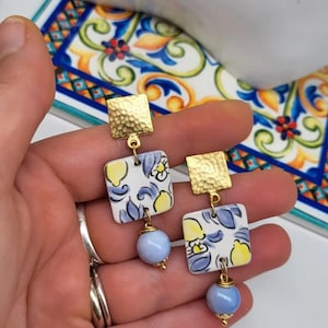 Può includere: Orecchini quadrati color oro con una texture martellata nella parte superiore. Gli orecchini presentano piastrelle quadrate in ceramica con un motivo a limoni e fiori e una perla azzurra nella parte inferiore. Un accessorio unico ed elegante.
