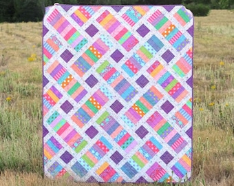 May include: A colourful quilt pattern called "The Iris Quilt" by Erica Jackman. The quilt is made with a variety of bright colours and patterns, including stripes, polka dots, and floral prints. The quilt is displayed in a field of green grass.