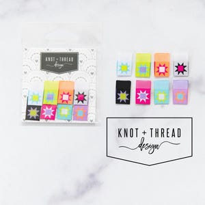 May include: A set of colorful fabric labels with starburst designs in a variety of colors, including yellow, orange, pink, and blue. The labels are packaged in a clear plastic bag, with the words "KNOT + THREAD design" printed on the packaging.