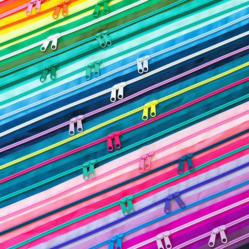 May include: A collection of nylon coil zippers arranged in a diagonal pattern, featuring a vibrant spectrum of colors including red, orange, yellow, green, blue, purple, and pink. Each zipper includes two matching plastic pull tabs.