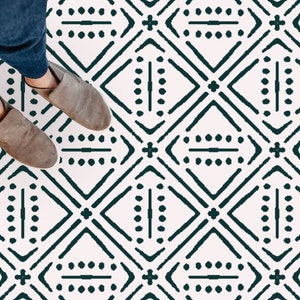 May include: A white floor with a dark green geometric pattern. The pattern is made up of diamonds, lines, and dots. A person is standing on the floor, wearing blue jeans and brown shoes.