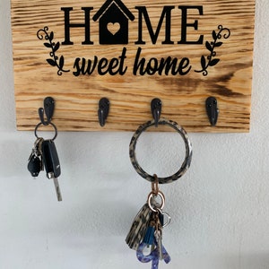 May include: Wooden key holder with the words "Home sweet home" and a house with a heart in the center. The key holder has four hooks and is decorated with a burnt wood finish. There are keys and a key chain with a tassel hanging from the hooks.