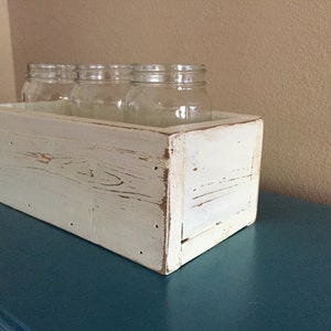 K&ouml;nnte beinhalten: Wei&szlig;e Holzkiste mit abgenutzter Farbe, die vier klare Glasgl&auml;ser enth&auml;lt. Die Kiste steht auf einer t&uuml;rkisfarbenen Oberfl&auml;che.