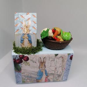 May include: A decorative box featuring illustrations of Peter Rabbit. The box has a light blue top and is adorned with images of carrots, cherries, and a basket of vegetables. The box is decorated with green moss.