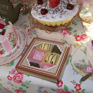 May include: A selection of baked goods on a table covered in a floral tablecloth. There are three cakes, one with pink and white checkered frosting and floral decorations, another with chocolate frosting and cherries, and a third with pink frosting and a floral design. There is also a box of small cakes and a plate with a single cake with pink frosting and a floral design.