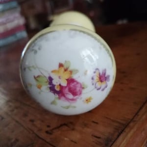 May include: A round porcelain door knob with a pale yellow base and a white centre featuring a painted floral arrangement of pink roses, yellow blossoms, and purple flowers. The edge of the knob is finished with a thin gold-coloured metallic trim.
