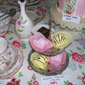 May include: A silver tiered cake stand with four fondant fancies, two with white and yellow stripes and two with pink frosting. The stand is on a white tablecloth with a floral pattern. A sign reads "Fondant Fancies 1.99 each."