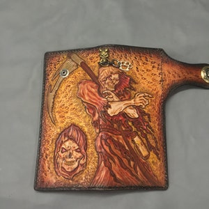 May include: Hand-tooled leather wallet featuring a Grim Reaper design. The wallet is primarily brown with orange and red accents, depicting the Reaper with a scythe and a skull. Includes a brass knuckle keychain.