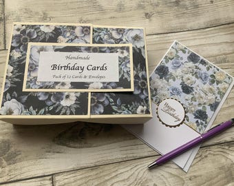 May include: A rectangular box with a dark blue and white floral pattern containing handmade birthday cards, with a label reading Handmade Birthday Cards Pack of 12 Cards & Envelopes, next to a floral greeting card and a purple ballpoint pen.