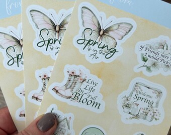 May include: A selection of spring-themed stickers with butterfly, boot, gnome, and floral designs. Text includes "Spring is in the Air", "Live Life in Full Bloom", and "Hello Spring". The stickers have a watercolour design with pastel colours.