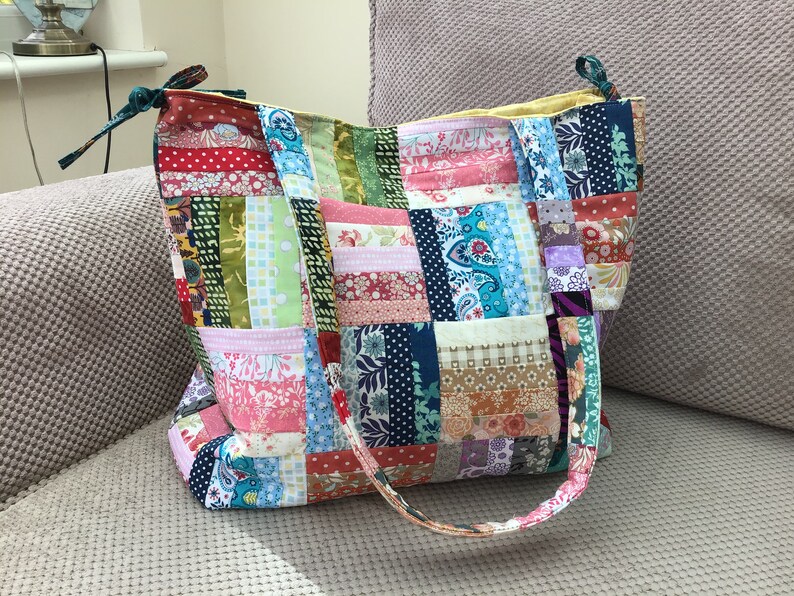 Peut inclure: Un sac fourre-tout en patchwork avec une vari&eacute;t&eacute; de tissus color&eacute;s, y compris des motifs floraux, paisley et g&eacute;om&eacute;triques. Le sac a deux longues lani&egrave;res et une fermeture &agrave; cordon.