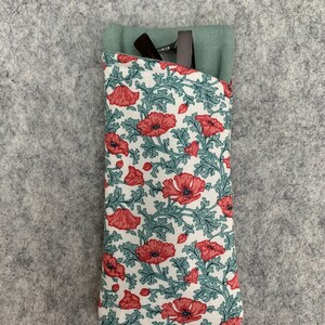 以下が含まれることがあります： 赤いポピーと緑の葉のフローラルパターンが施された白い布製の眼鏡ケース。ケースは折り畳んで閉じられており、上部に緑色の布製のフラップが付いています。