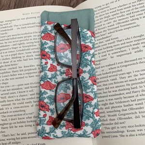 以下が含まれることがあります： 本のアップで、花柄のしおりと老眼鏡が置かれています。本は「結婚式」と「クラム」というキャラクターについてのテキストのあるページを開いています。