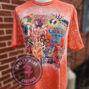 Peut inclure: T-shirt orange d&eacute;lav&eacute;e avec un imprim&eacute; graphique de Janis Joplin, Woodstock et d'autres ic&ocirc;nes de la musique et de la culture des ann&eacute;es 1960. Le t-shirt a un aspect vieilli avec des &eacute;claboussures de peinture blanche.