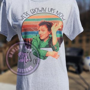 May include: A gray t-shirt with a retro-style graphic of a man with a green jacket and the text "We're Grown Ups Now".