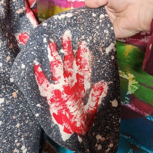 May include: Dark gray sweatshirt sleeve with a large red handprint design. The fabric has a speckled pattern with white and tan spots. The sleeve is part of a larger garment.