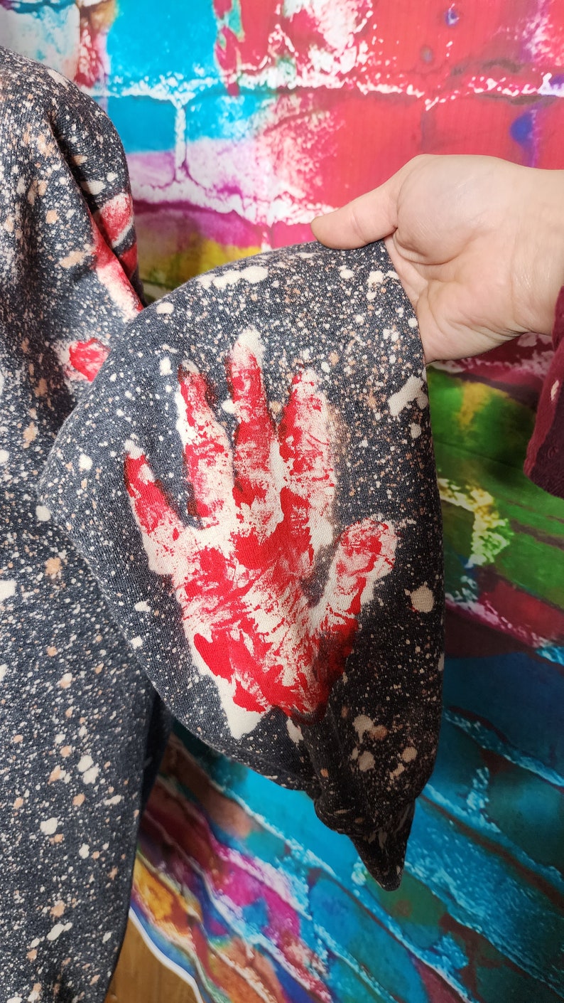 May include: Dark gray sweatshirt sleeve with a large red handprint design. The fabric has a speckled pattern with white and tan spots. The sleeve is part of a larger garment.