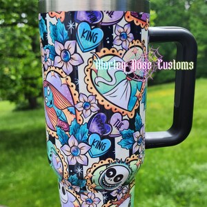 May include: A black and silver travel mug with a black handle and lid. The mug is decorated with a colorful pattern of flowers, hearts, and characters from The Nightmare Before Christmas. The text "KING" is visible on the mug.