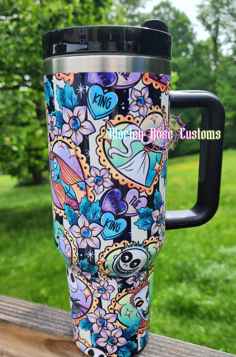 May include: A black and silver travel mug with a black handle and lid. The mug is decorated with a colorful pattern of flowers, hearts, and characters from The Nightmare Before Christmas. The text "KING" is visible on the mug.