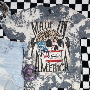May include: Gray t-shirt with a bleached effect featuring a skull with an American flag design and leopard print. The text "Made in America" is printed on the shirt.
