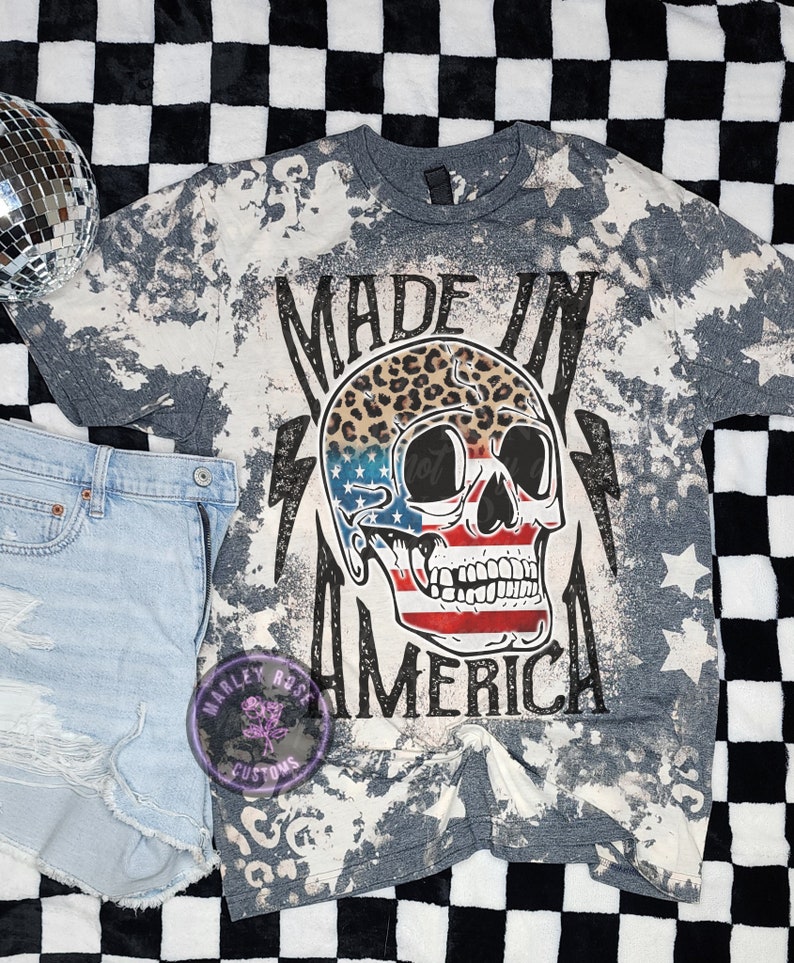 May include: Gray t-shirt with a bleached effect featuring a skull with an American flag design and leopard print. The text "Made in America" is printed on the shirt.