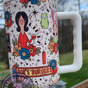 May include: A white ceramic mug with a black handle and the text "But First, Coffee" in a black, bold font. The mug is on a white surface. The mug is approximately 4 inches tall.