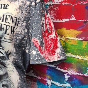 May include: Dark gray t-shirt with a distressed newspaper print design and white splatter effect. The sleeve features a large red handprint. The shirt has a vintage, grunge aesthetic.