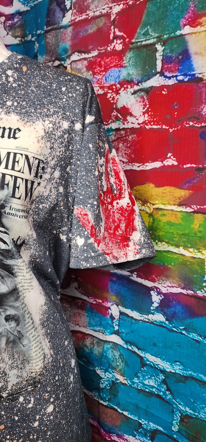 May include: Dark gray t-shirt with a distressed newspaper print design and white splatter effect. The sleeve features a large red handprint. The shirt has a vintage, grunge aesthetic.