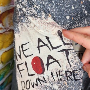 May include: A gray and white t-shirt with the text "WE ALL FLOAT DOWN HERE" and a red balloon graphic. The shirt has a distressed, speckled design.