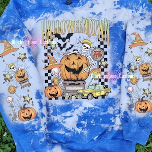 May include: A blue and white tie-dye sweatshirt featuring a graphic print with the text HALLOWEENTOWN EST 1998. The design includes a skeleton, a large jack-o'-lantern, a yellow taxi, bats, spiderwebs, witch hats, and lollipops on the front and sleeves.