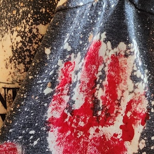 May include: Dark gray t-shirt sleeve with a large red handprint and white splatter design. The fabric has a speckled pattern, and the handprint is prominently displayed. The overall aesthetic is edgy and artistic.