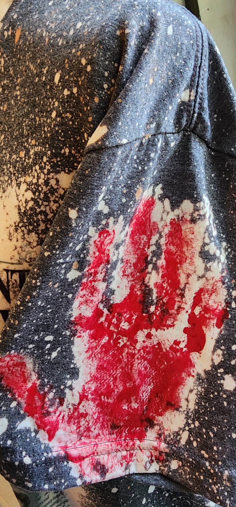 May include: Dark gray t-shirt sleeve with a large red handprint and white splatter design. The fabric has a speckled pattern, and the handprint is prominently displayed. The overall aesthetic is edgy and artistic.