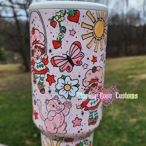 May include: A pink tumbler with a white straw, featuring a Strawberry Shortcake theme. The design includes illustrations of Strawberry Shortcake, butterflies, strawberries, flowers, and suns. The text "Morley Rose Customs" is also visible.