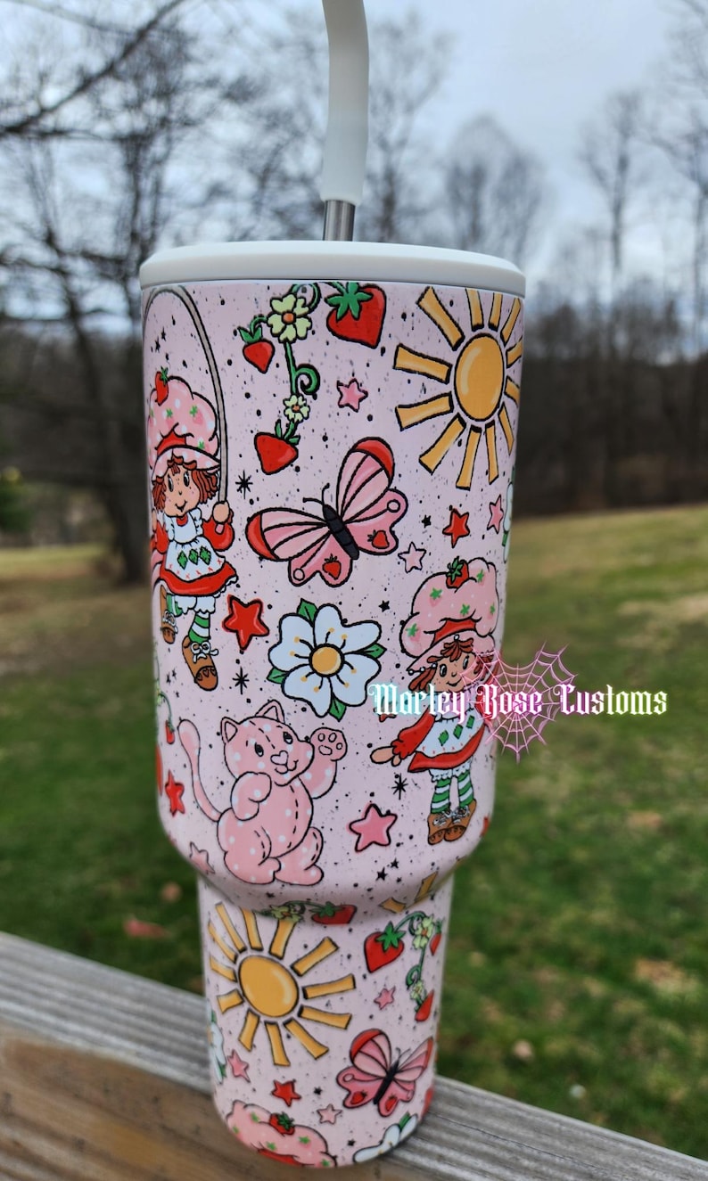 May include: A pink tumbler with a white straw, featuring a Strawberry Shortcake theme. The design includes illustrations of Strawberry Shortcake, butterflies, strawberries, flowers, and suns. The text "Morley Rose Customs" is also visible.