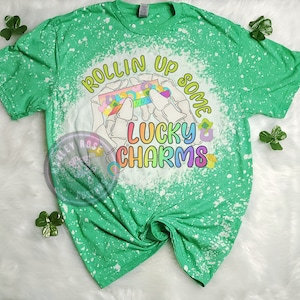 May include: Green t-shirt with a bleach-splattered design featuring a hand holding a box of Lucky Charms cereal with the text "Rollin' Up Some Lucky Charms".