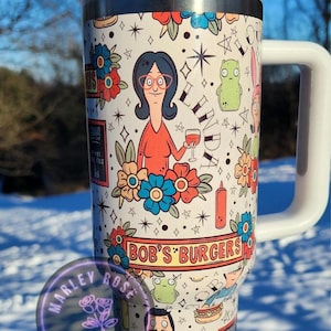 May include: A white travel mug featuring characters and imagery from the animated series "Bob's Burgers." The design includes floral accents, cartoon figures, and the show's logo. The mug has a white handle and lid.