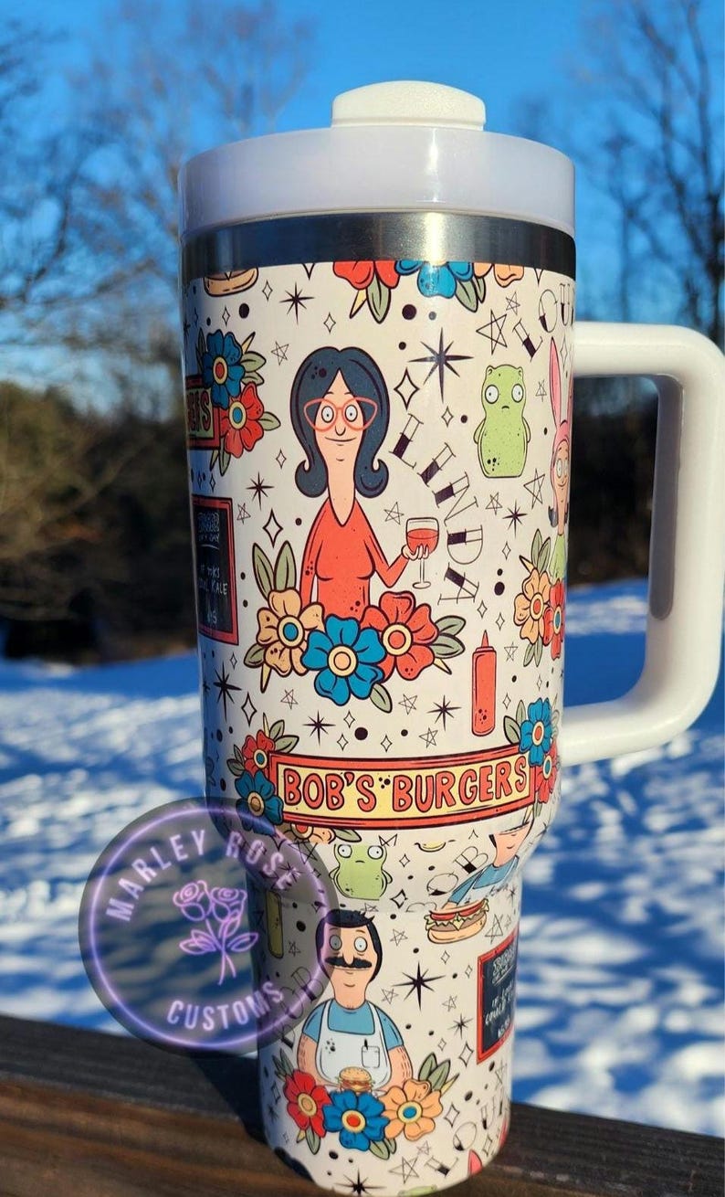 May include: A white travel mug featuring characters and imagery from the animated series "Bob's Burgers." The design includes floral accents, cartoon figures, and the show's logo. The mug has a white handle and lid.