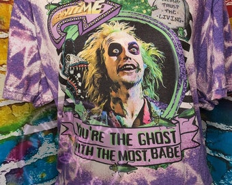 May include: Purple tie-dye t-shirt featuring a graphic of a character with green hair and the text "YOU'RE THE GHOST WITH THE MOST, BABE." The design includes the words "WELCOME" and "NEVER TRUST THE LIVING."