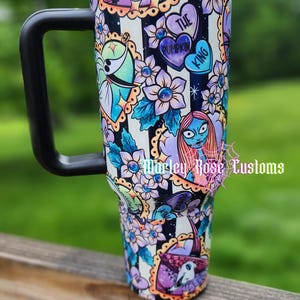 May include: A stainless steel travel mug with a black handle and lid. The mug is decorated with a colorful design featuring characters and floral elements from "The Nightmare Before Christmas" against a black and white striped background.