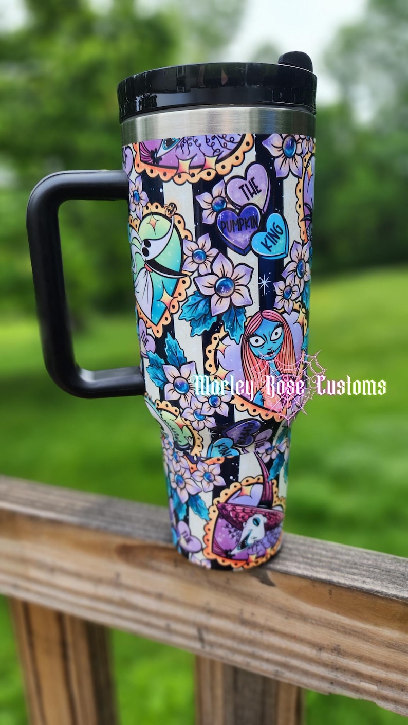 May include: A stainless steel travel mug with a black handle and lid. The mug is decorated with a colorful design featuring characters and floral elements from "The Nightmare Before Christmas" against a black and white striped background.