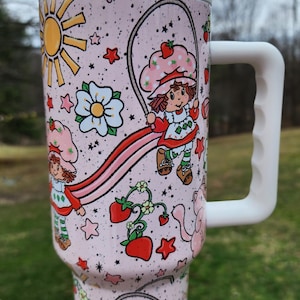 May include: A pink tumbler with a white handle and straw, featuring a cartoon design of a girl with a strawberry hat, jumping rope. The design includes strawberries, stars, and a sun. The text "Marley Rose Customs" is visible.